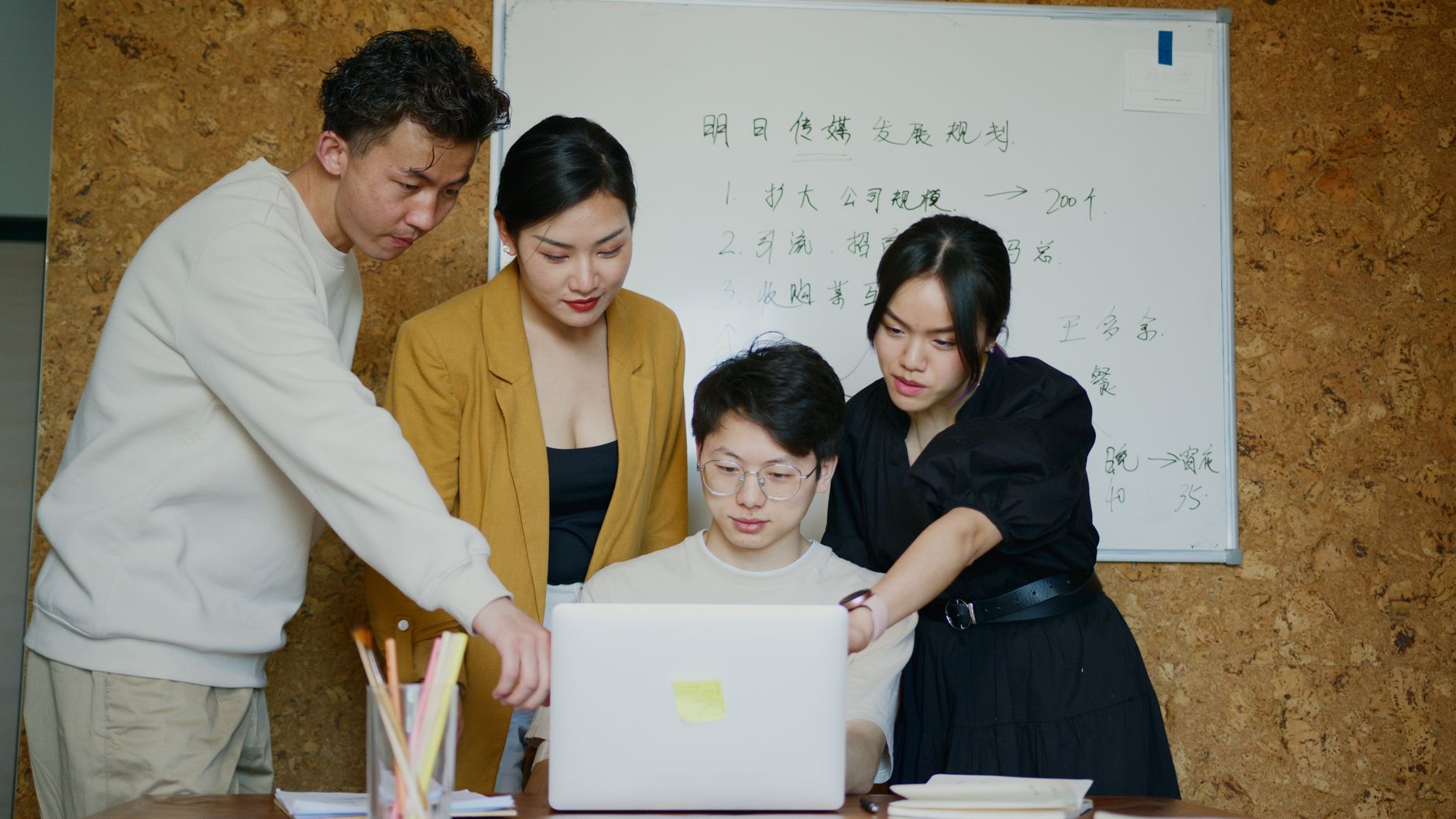 In an open office, one developer types rapidly alone while teammates gather at a whiteboard behind them, debating a complex diagram